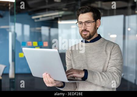 Professional man wearing eyeglasses standing and working on a laptop computer in a contemporary office setting, with glass walls and colorful sticky notes in the background Stock Photo