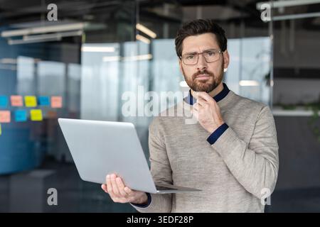 Thoughtful businessman with a beard and glasses holding a laptop, standing in a contemporary office setting while pondering solutions, problems or new ideas, indicating deep contemplation Stock Photo