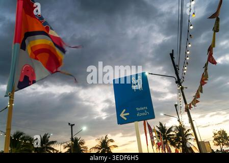 Sunset illuminates a colorful flag and a blue toilet sign amidst cloudy skies, creating a vibrant atmosphere in a tropical outdoor setting. Stock Photo