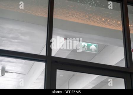 A view of an emergency exit sign visible through a large window in a modern building. The glowing green sign emphasizes safety and direction in indoor Stock Photo