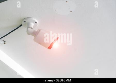 A sleek security camera mounted on the ceiling emits a red light, showcasing modern surveillance technology in an indoor setting for enhanced safety a Stock Photo