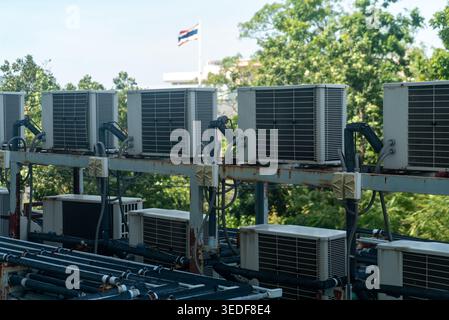 A row of air conditioning units is installed on a building rooftop, showcasing modern cooling technology against a backdrop of trees and a flag. Stock Photo