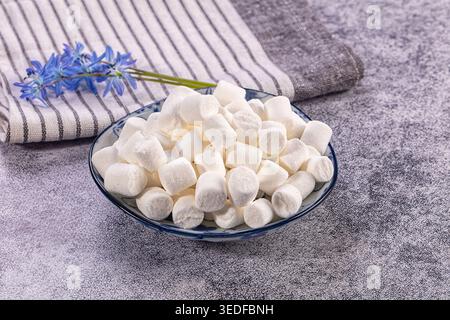Sweet white mini marshmallows piled in decorative ceramic bowl. Fluffy treats sit beside striped linen napkin, delicate blue spring flowers Stock Photo