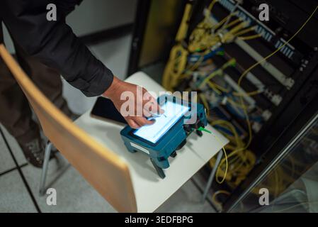 A technician operates a handheld device in a data center. The photo captures a close-up of the technician’s hand using a touchscreen for network analy Stock Photo