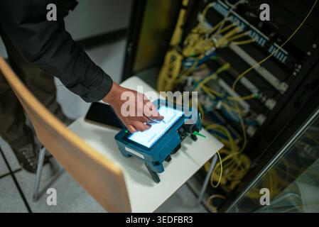 A technician is using a handheld device to diagnose network issues in a server room, highlighting modern technology in action amid cables and hardware Stock Photo
