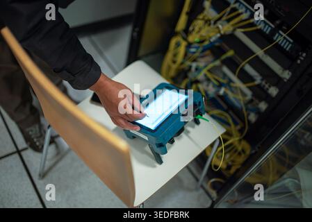A technician operates a portable device in a modern data center, focusing on network analysis and monitoring, surrounded by network cables and equipme Stock Photo