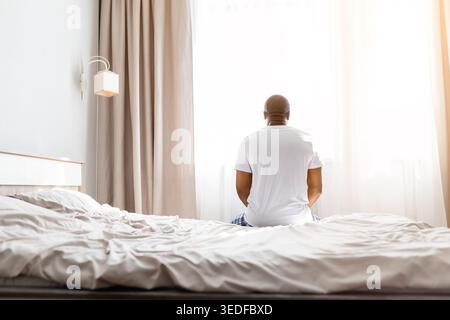 Black man sitting on edge of unmade bed in pajamas, rear view Stock Photo