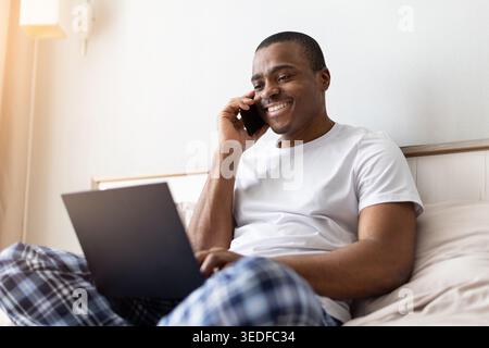 African american man talking on smartphone while using laptop on bed Stock Photo