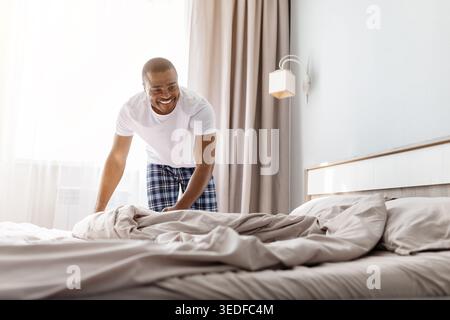 Happy African American man smiling while making bed in bright bedroom Stock Photo