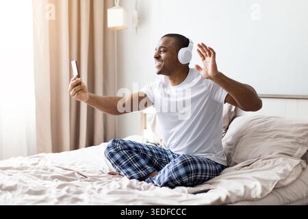 Smiling african american man wearing headphones waving at smartphone on bed Stock Photo
