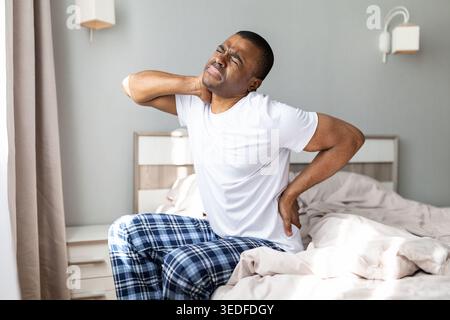 African american man experiencing neck and back pain while sitting on bed Stock Photo