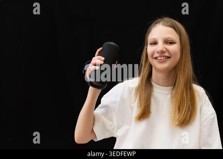 Smiling Teen Girl Holding Portable Speaker. Happy blonde teen girl holding a black wireless speaker, representing music, audio technology Stock Photo