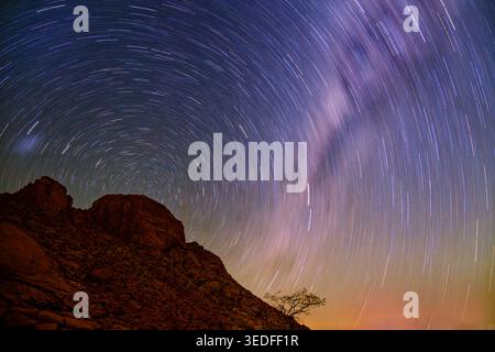 Star trails over Spitzkoppe granite inselberg, Namibia, long exposure night sky, circular star rotation, Milky Way, lone tree silhouette, desert lands Stock Photo