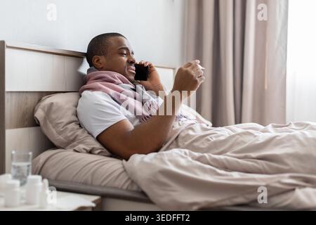Black man lying in bed talking on phone while checking thermometer Stock Photo