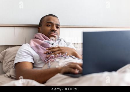 Sick black man sitting in bed with blanket and scarf, using laptop Stock Photo