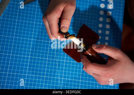 Artisan in a leather apron preparing an edge creasing tool with a lighter over a blue mat Stock Photo