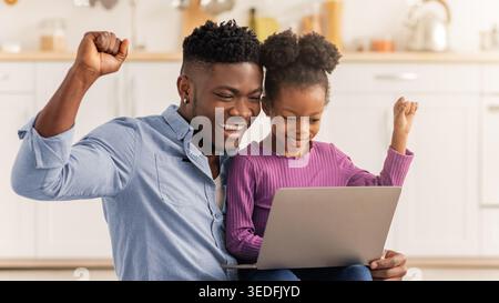 Father and Daughter Celebrate Success While Using a Laptop in a Bright Kitchen Stock Photo