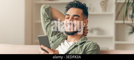 Man Relaxes on Couch While Using Smartphone and Listening to Music With Headphones Stock Photo