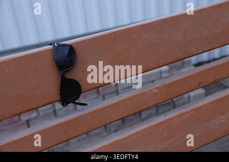 Sunglasses rest on the slats of a wooden bench. The scene shows a simple outdoor setting near a wall. The sun is bright and creates a casual atmosphere Stock Photo