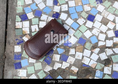 A brown leather case sits on mosaic tiles made of small pieces of colored stone. The setting is outdoors with sunlight shining down. The tiles are arranged in various colors Stock Photo