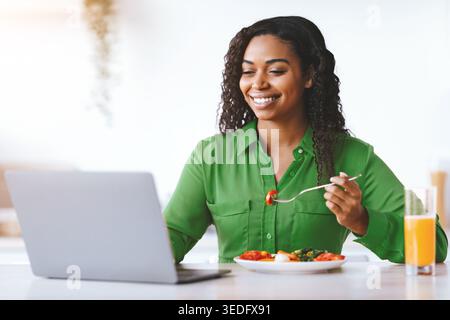 Young Woman Enjoys Meal While Working on Laptop in Bright Indoor Space During Day Stock Photo