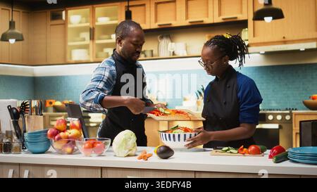 Black couple in aprons adding vegetables in a bowl and mixing ingredients, making a fresh vegetarian salad for their lunch meal. Healthy lifestyle and positive relationship. Stock Photo