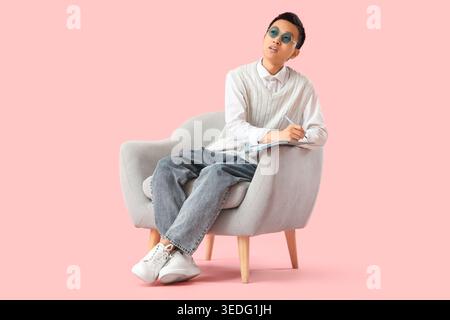 Thoughtful young Asian man writing something in notebook on armchair against pink background Stock Photo