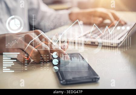 A business professional uses a smartphone to analyze financial data while working on a laptop, with graphs and charts visually displaying performance Stock Photo