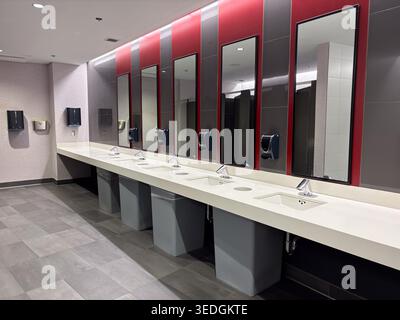 A clean, modern men's restroom featuring a long vanity with multiple sinks, soap dispensers, and wall mirrors against a red and gray tiled wall. Stock Photo