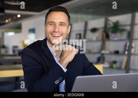 Smiling businessman sits at office desk, laughing, working on laptop Stock Photo