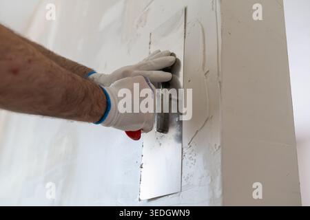 Application of a finishing coat using a metal trowel on drywall in residential renovation. The worker wears protective gloves for safety while smoothing the compound. Stock Photo