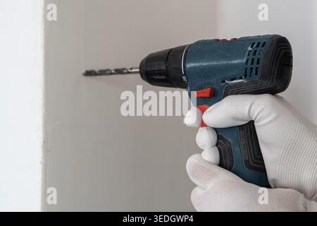 Electric drill in use, applying pressure against wall surface for drilling. Gloved hand holds tool firmly while working in a clean indoor environment. Stock Photo