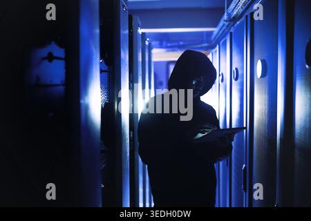 Hacker, tablet and criminal with typing in server room for cyber crime, phishing or programming. Person, tech and hoodie for malware, application or Stock Photo
