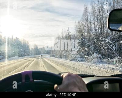 A car drives along a snowy road surrounded by trees on a winter day. The sun shines brightly through the clouds. Stock Photo