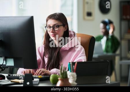 Portrait of young adult Caucasian woman working at desktop computer in office, looking at camera and typing. Suitable for remote work, startup team, productivity marketing Stock Photo