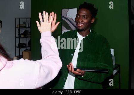 African American young adult man giving high five while holding laptop during office meeting, showing teamwork and success. Useful for business collaboration, onboarding, workplace culture Stock Photo