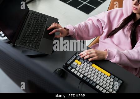 Young adult woman working on laptop and external keyboard at desk, managing digital tasks and multitasking in home office. Ideal for remote work, productivity, tech workflow Stock Photo
