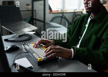 African American young adult man typing on keyboard while wearing headphones at home workstation, working remotely on laptop. Ideal for remote work, productivity, freelance content Stock Photo