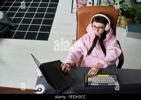 Young adult Caucasian woman wearing headphones, working on laptop and keyboard at home office, multitasking in remote workflow. Useful for hybrid work, freelance, productivity marketing Stock Photo