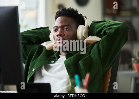 Medium shot of young African American man wearing headphones, leaning back while working at computer, taking break in home office. Ideal for remote work, focus, productivity content Stock Photo