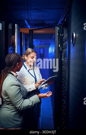 Women, tablet and team with review in server room with cybersecurity, system check or collaboration. Programmer, tech and advice with smile for Stock Photo