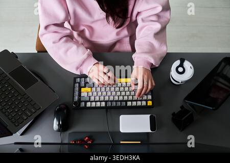 Young adult woman typing on keyboard at desk with laptop, smartphone and coffee, working in home office setup. Top view, fitting remote work, productivity, freelance marketing use Stock Photo