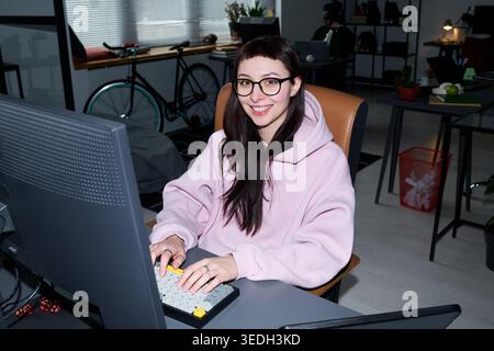 Portrait of young adult Caucasian woman working at desktop computer, typing in office and looking at camera. Ideal for remote work, startup, productivity, tech career marketing Stock Photo