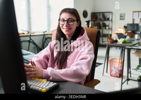 Portrait of young adult Caucasian woman working at desktop computer in office, smiling at camera. Ideal for remote work, startup team, productivity, career content Stock Photo