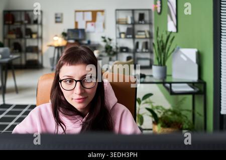 Young adult Caucasian woman working at computer in home office, focusing on screen during remote task. Ideal for remote work, freelance business, productivity marketing, medium shot Stock Photo