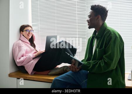 Young adult Caucasian woman using laptop while talking with African American man holding tablet, collaborating in casual workspace. Ideal for remote work, startup teamwork, digital communication Stock Photo