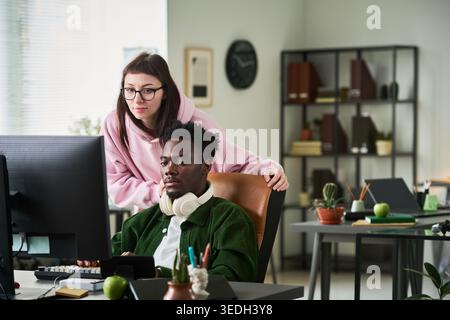 Young Black man working at desktop computer while Caucasian woman guiding project review in office, showing teamwork and mentoring. Ideal for workplace collaboration campaigns Stock Photo
