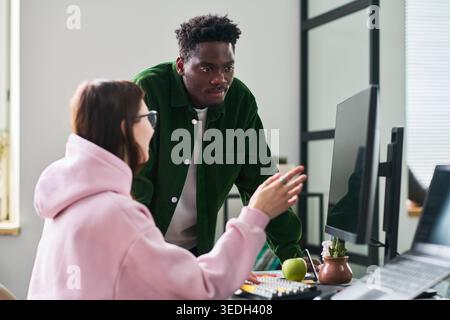 Young adult African American man and Caucasian woman collaborating at computer, reviewing project and discussing workflow. Ideal for teamwork, office communication, creative agency Stock Photo