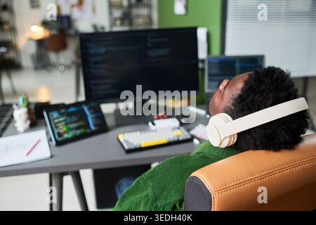 African American young adult man leaning back at workstation, wearing headphones, reviewing code on multiple screens and tablet, representing remote software work. For tech hiring or productivity Stock Photo