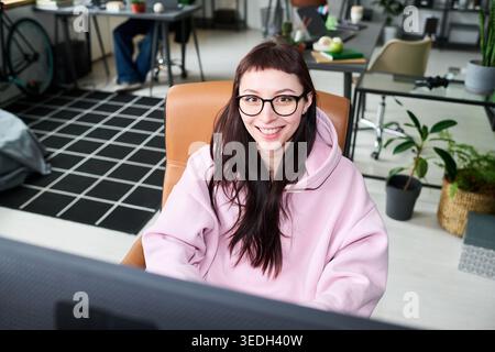 Portrait of young adult Caucasian woman wearing glasses, sitting at computer in modern office, smiling at camera. Ideal for remote work, startup, digital career marketing Stock Photo